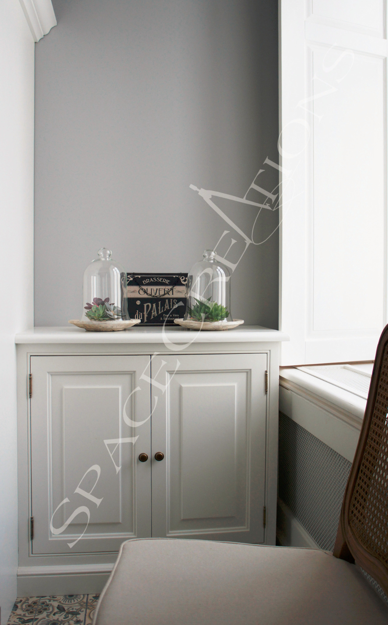 Szara szafka z dekoracjami (szklane klosze z sukulentami, tabliczka 'Brasserie Ouvert') w rogu pokoju z krzesłem. Aranżacja wnętrza w stonowanej kolorystyce.