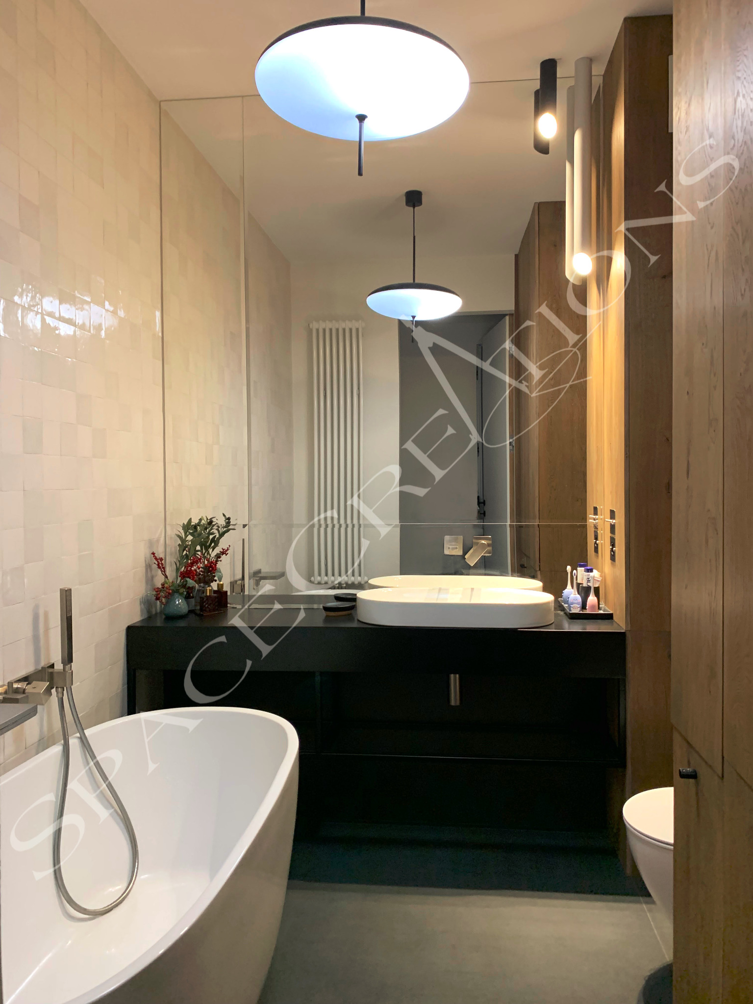 Nowoczesna łazienka z wolnostojącą wanną, umywalką nablatową i dużym lustrem. Ściany wyłożone jasnymi, kwadratowymi płytkami, oświetlenie punktowe i wiszące.