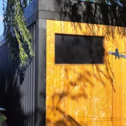 Nowoczesna altana ogrodowa z drewna, z oknem, pomalowana na czarno i żółto, oświetlona słońcem z cieniem drzewa. Widok z boku, z fragmentem roślinności.