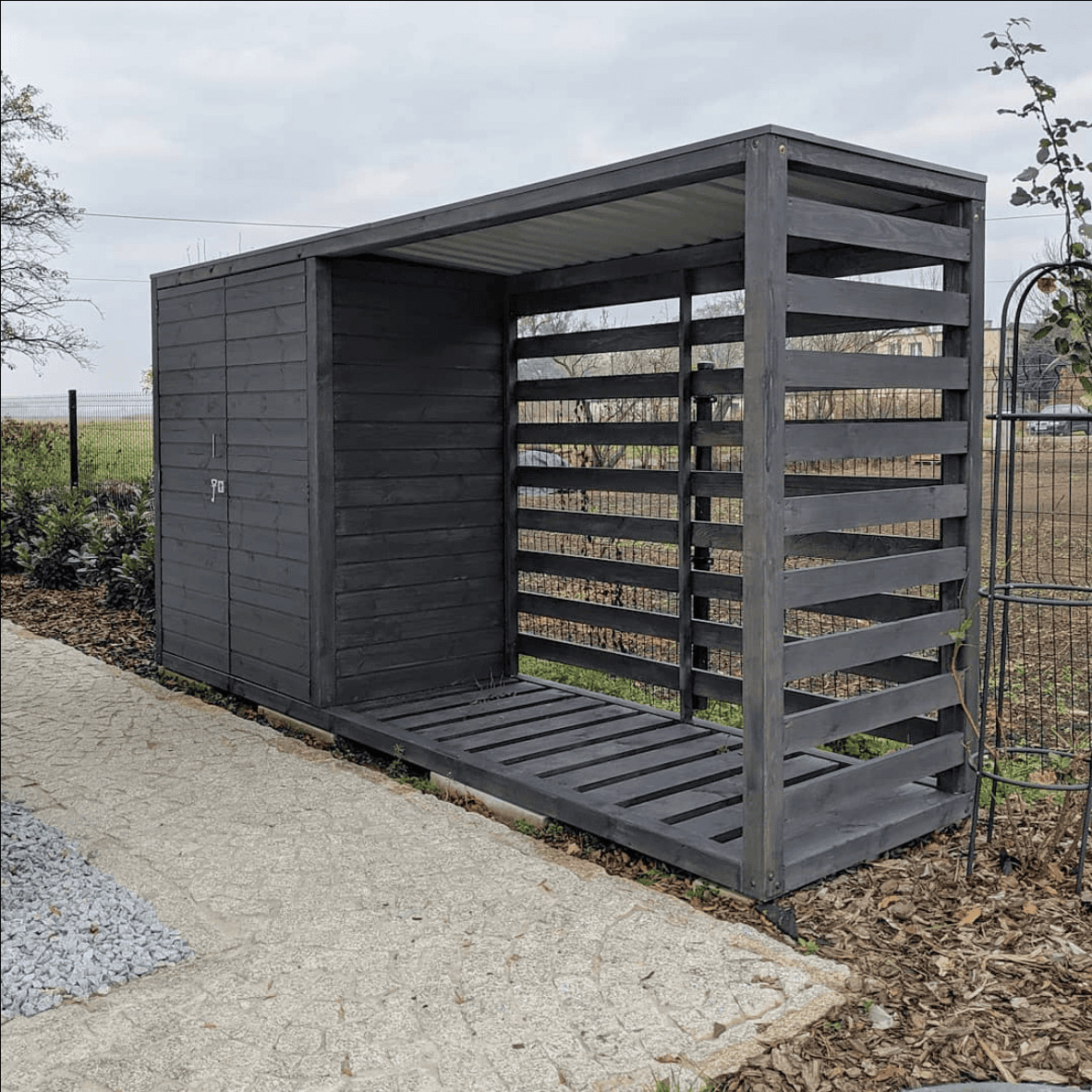 Nowoczesna drewutnia z szarego drewna z zamykaną częścią i otwartą przestrzenią na drewno, z metalowym dachem, na tle ogrodu.