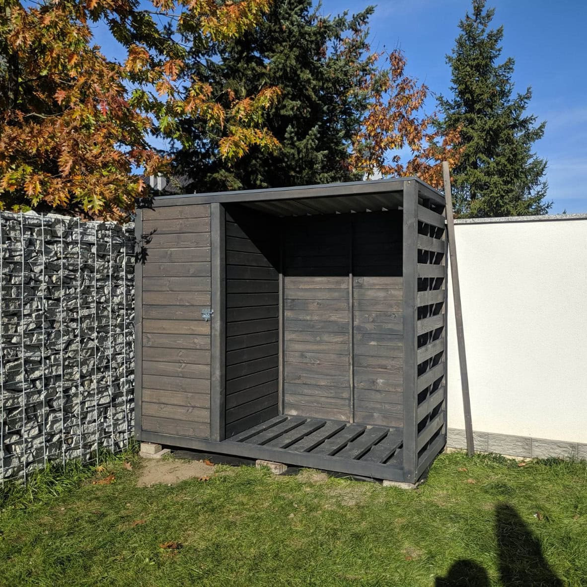 Nowoczesna, ciemna drewutnia z drewna w Dobra, zamykana na zamek, z ażurowymi ścianami, na trawie, obok gabionu i białego muru, jesienne drzewa w tle.