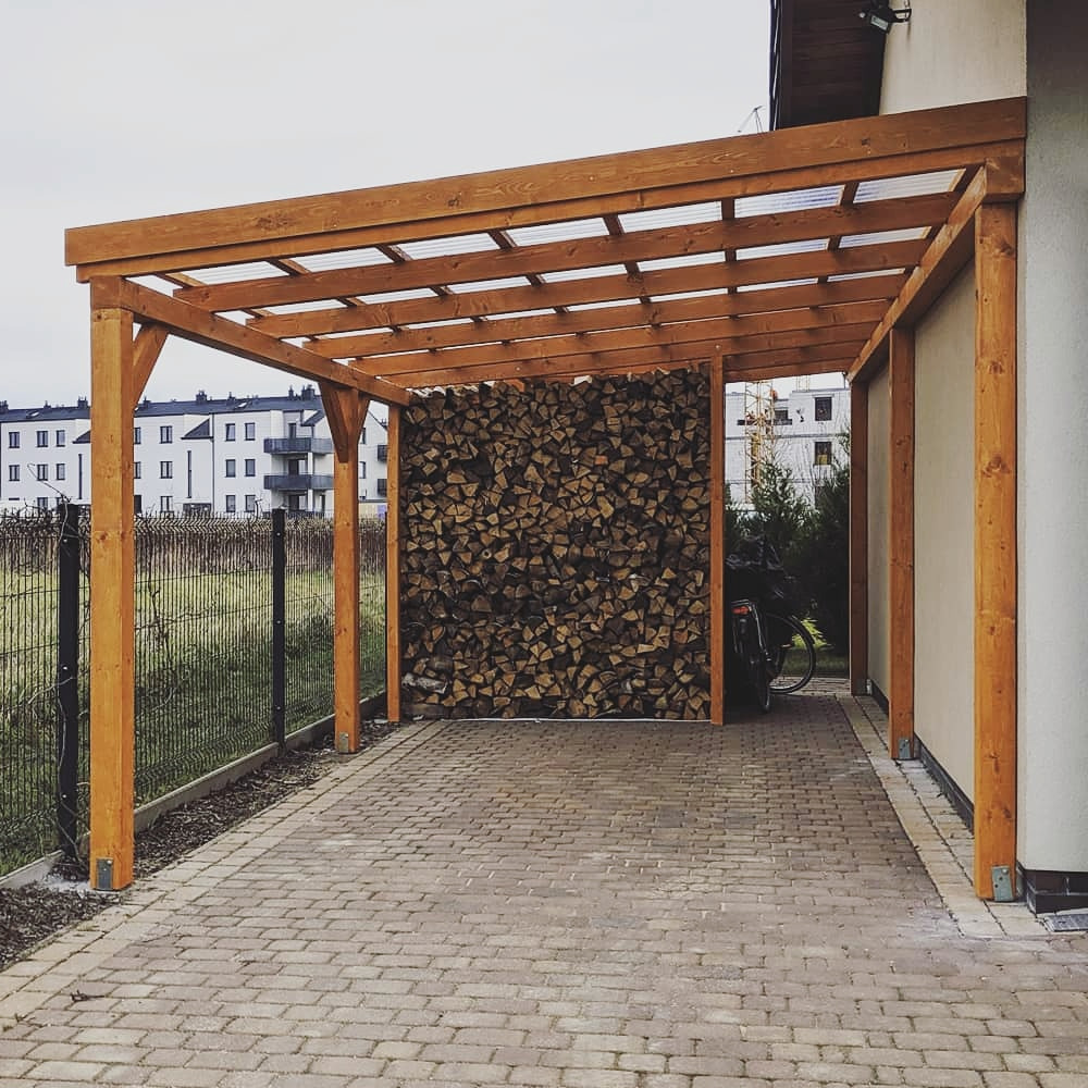 Nowoczesna wiata garażowa z drewna w minimalistycznym stylu, z transparentnym zadaszeniem. Na ścianie ułożone drewno opałowe. Brukowana podłoga.