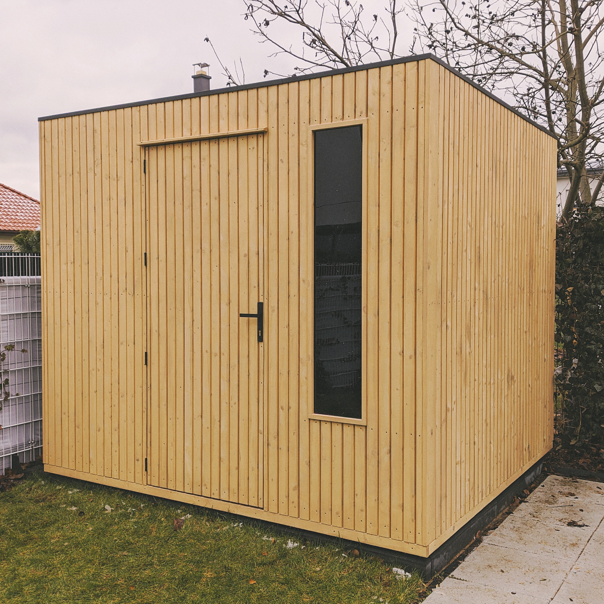 Minimalistyczny, nowoczesny domek ogrodowy z jasnego drewna z pionowymi deskami, czarnym oknem i drzwiami, na trawniku obok betonowej ścieżki.