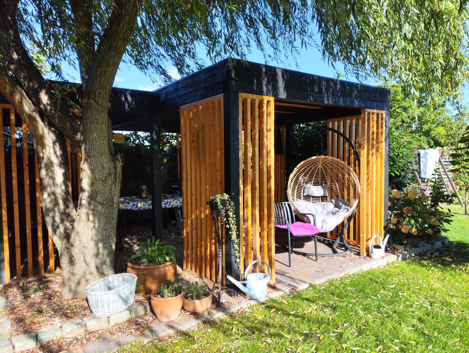 Nowoczesna altana ogrodowa z czarną konstrukcją i drewnianymi lamelami, z wiszącym fotelem i zielenią w tle, oświetlona naturalnym światłem słonecznym.