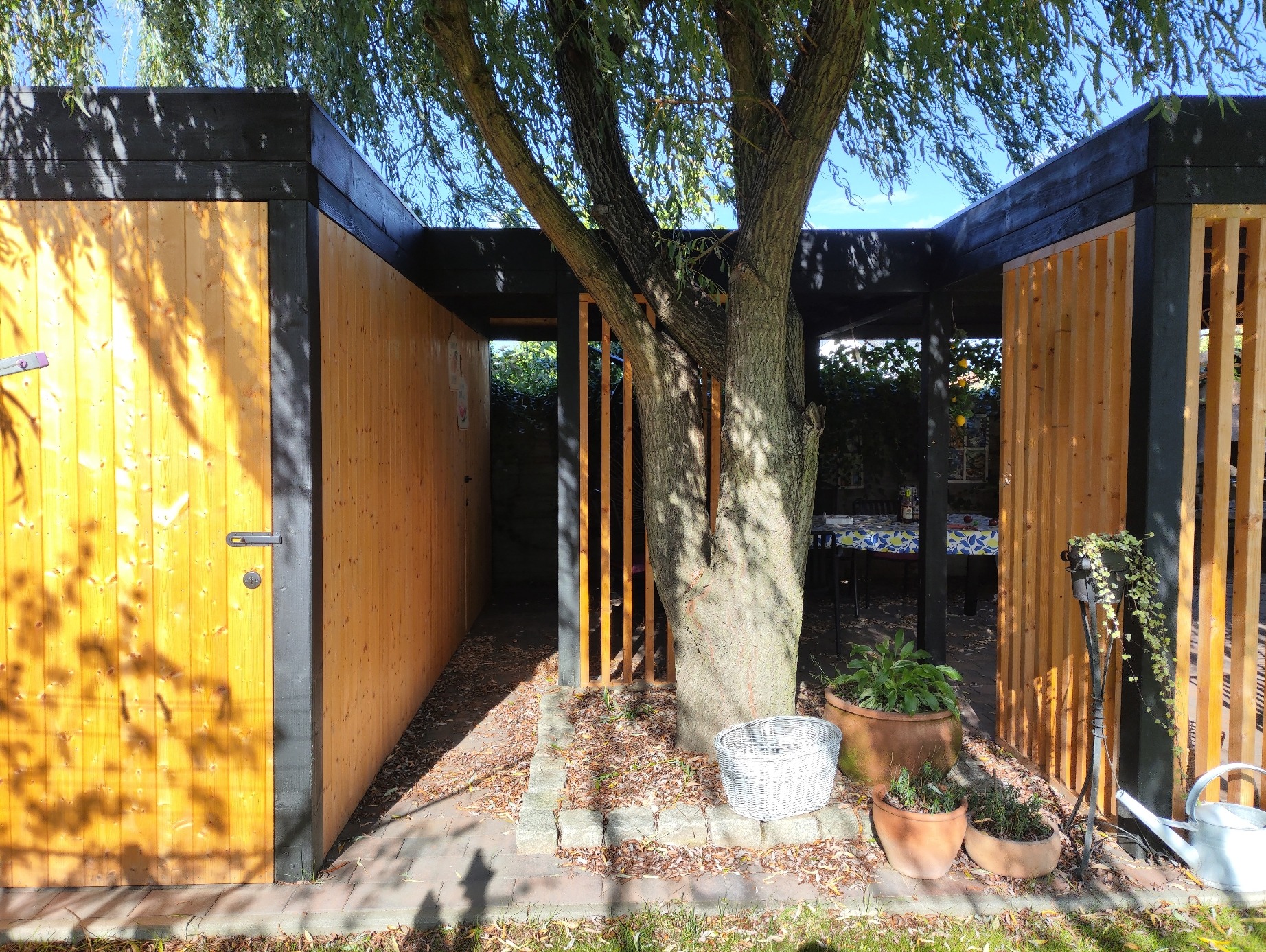 Nowoczesna altana ogrodowa z drewna w kolorze naturalnym i czarnym, częściowo zabudowana, z drzewem w centrum. Widoczne donice i elementy wyposażenia.