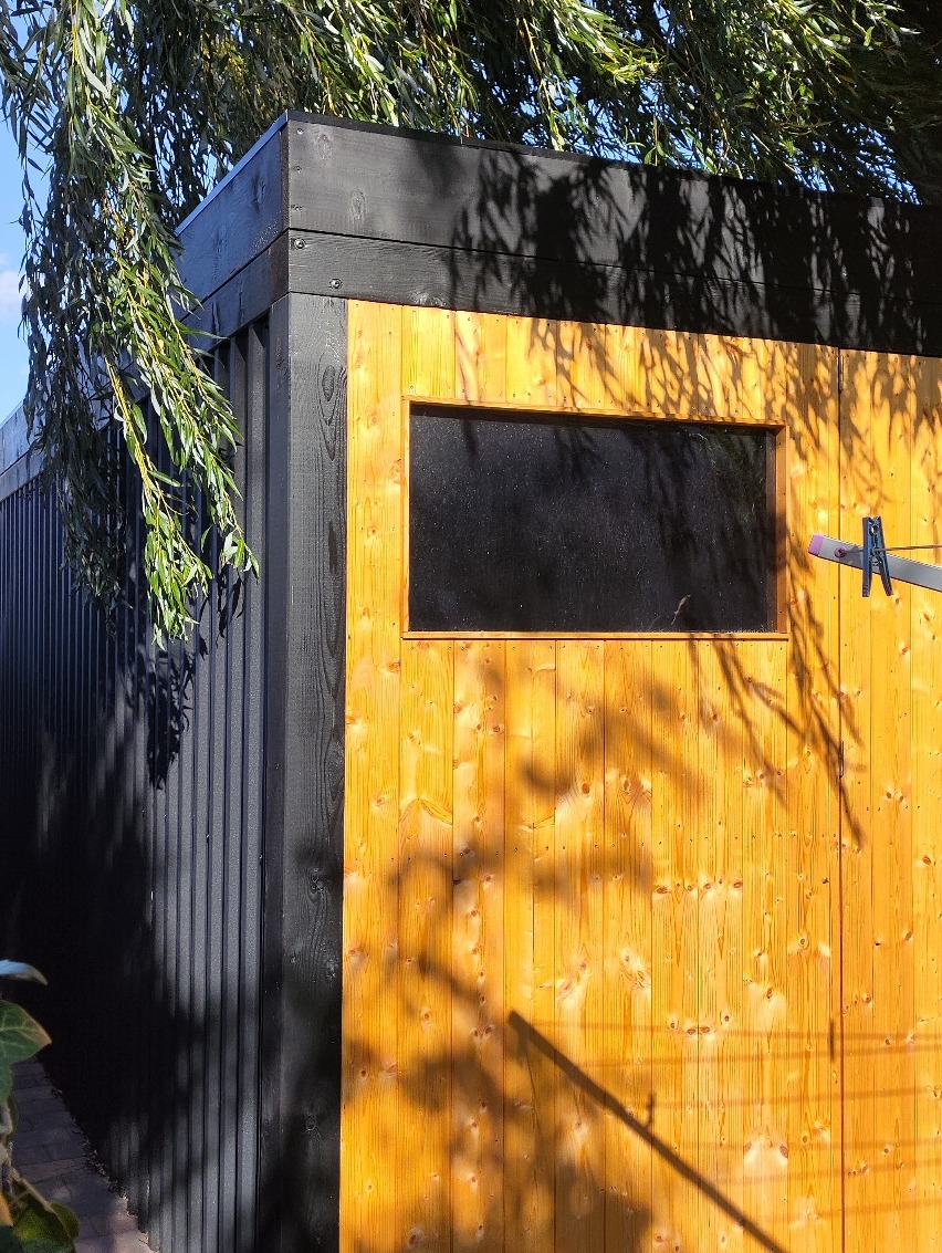 Nowoczesna altana ogrodowa z drewna, z oknem, pomalowana na czarno i żółto, oświetlona słońcem z cieniem drzewa. Widok z boku, z fragmentem roślinności.