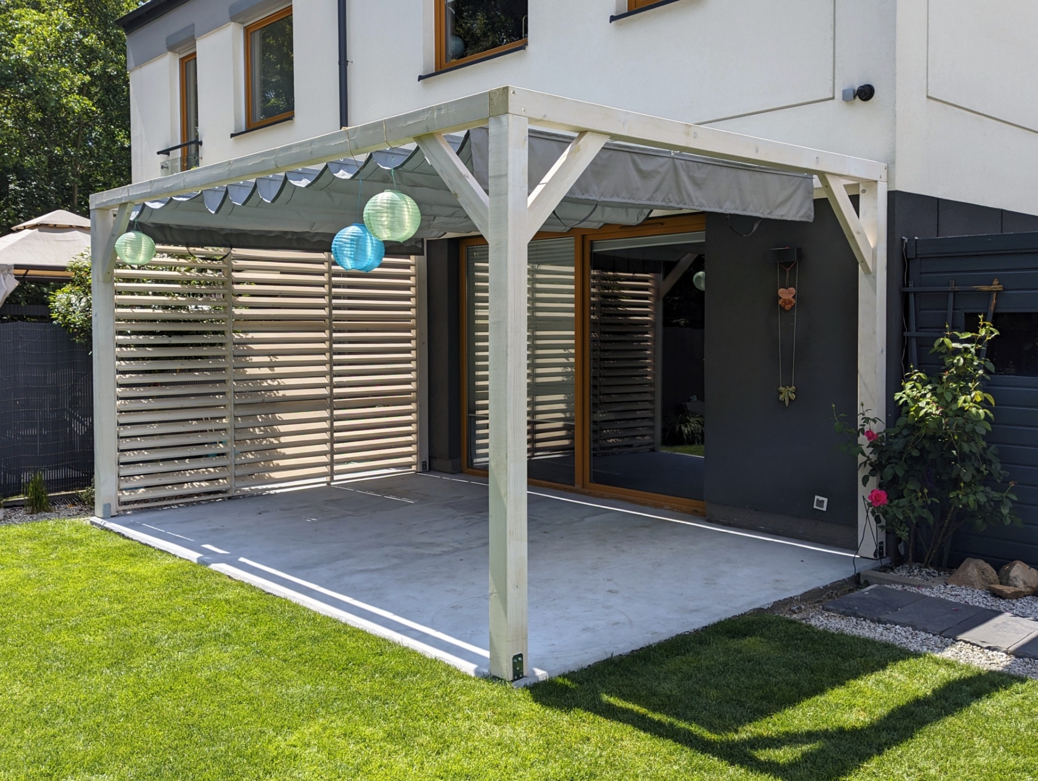 Nowoczesna pergola z drewnianą ścianką i szarą zasłoną, ozdobiona lampionami, przylegająca do budynku z wyjściem na taras i trawnik.
