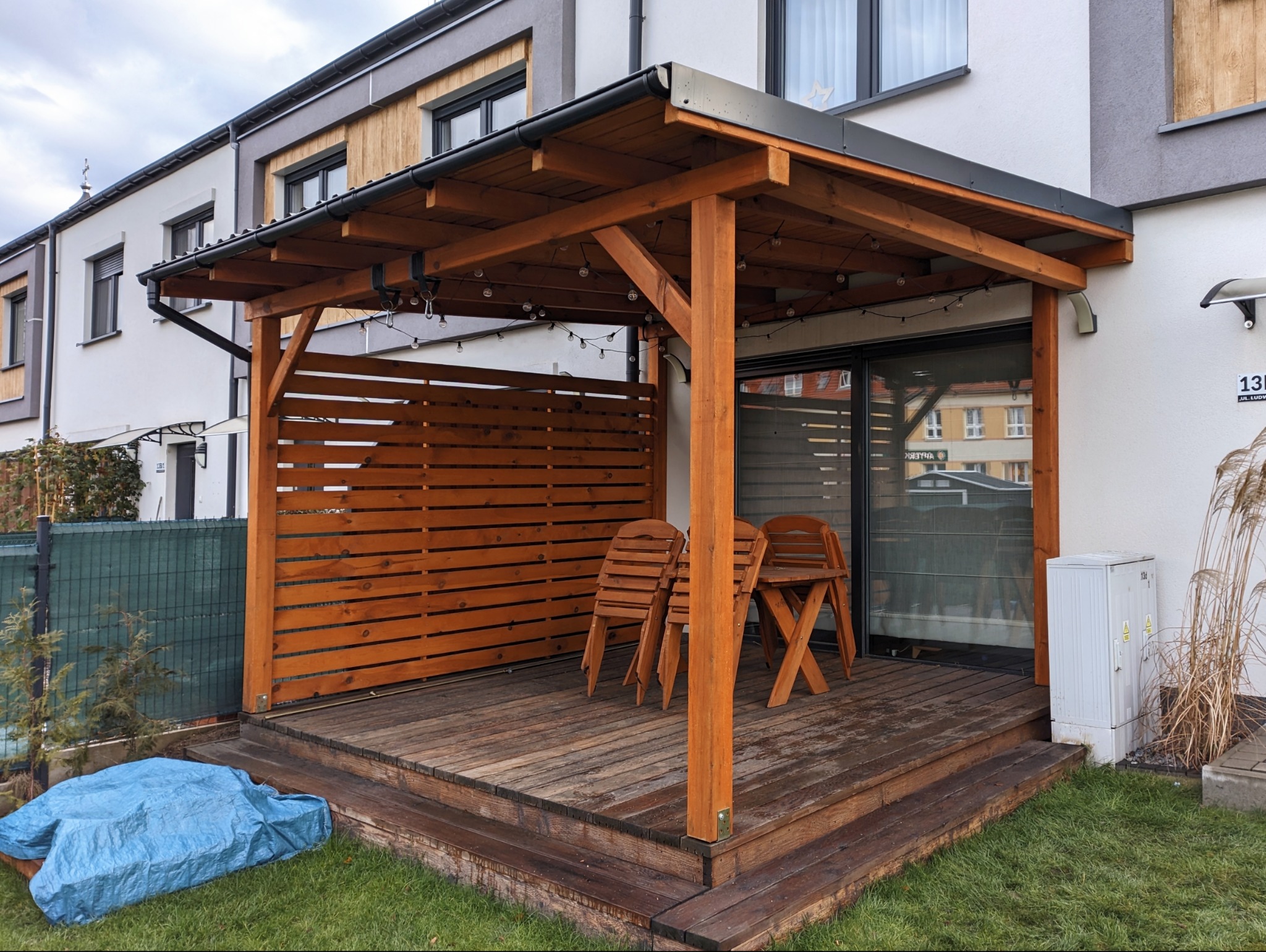 Klasyczna altana ogrodowa z drewnianym tarasem i ażurową ścianą, oświetlona girlandą. Na tarasie złożone krzesła. Obok budynku skrzynka elektryczna i trawa.