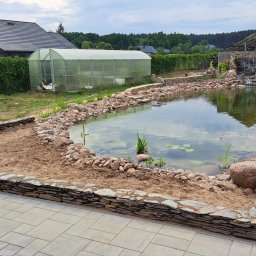 Ogrody Skalne - Oczko wodne w trakcie budowy z kamienną kaskadą i roślinami wodnymi, otoczone kamieniami i piaskiem. W tle szklarnia i dom z polską flagą.