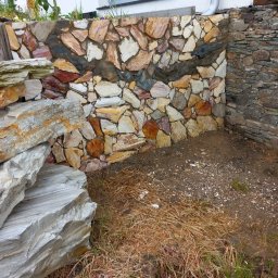 Ogrody Skalne - Kamienny murek oporowy z nieregularnych, kolorowych płyt. Widoczne fragmenty ogrodu i elewacji budynku. Wykończenie z kamienia w odcieniach beżu, brązu i szarości.