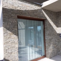 Ogrody Skalne - Ściana zewnętrzna wyłożona dekoracyjnym kamieniem elewacyjnym w odcieniach szarości i beżu, z oknem w brązowej ramie. Widoczne fragmenty tarasu i zadaszenia.