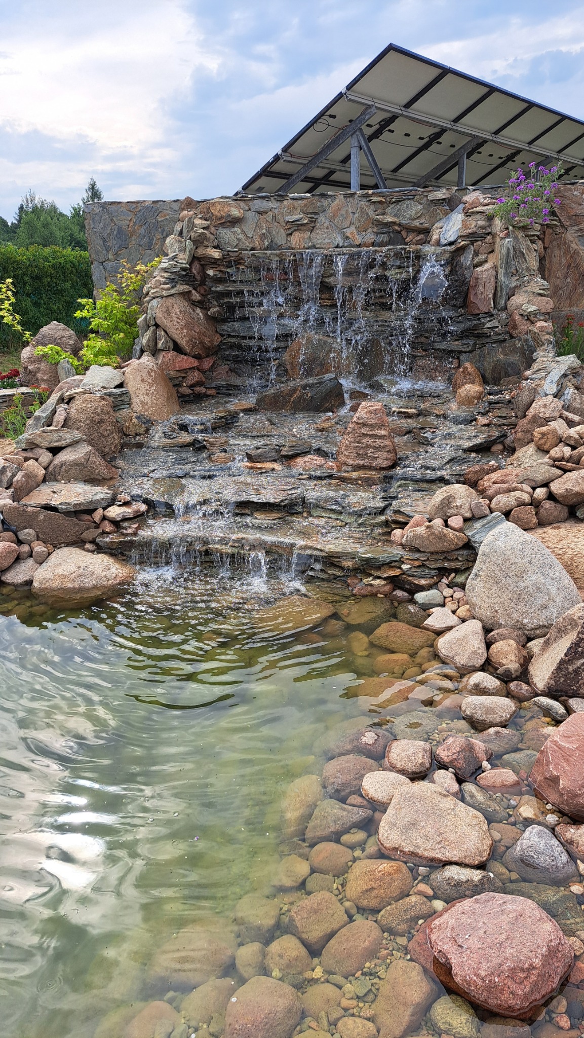 Kaskada wodna z kamienia w ogrodzie, zasilana energią słoneczną. Woda spływa po skałach do oczka wodnego z widocznymi kamieniami na dnie. W tle konstrukcja z panelami słonecznymi.