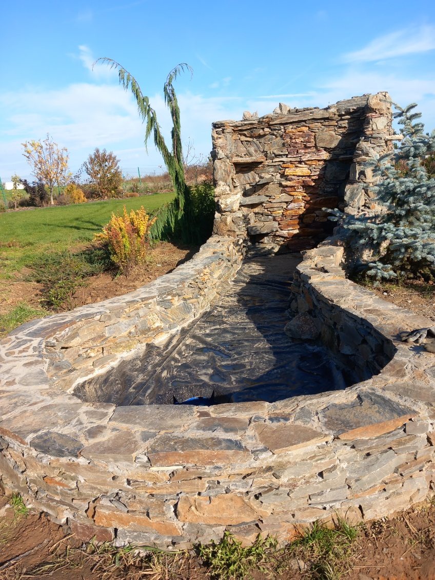 Oczko wodne w ogrodzie, wyłożone kamieniem, z wodospadem z kamienia. Widoczna folia zabezpieczająca. W tle trawnik i drzewa pod błękitnym niebem.