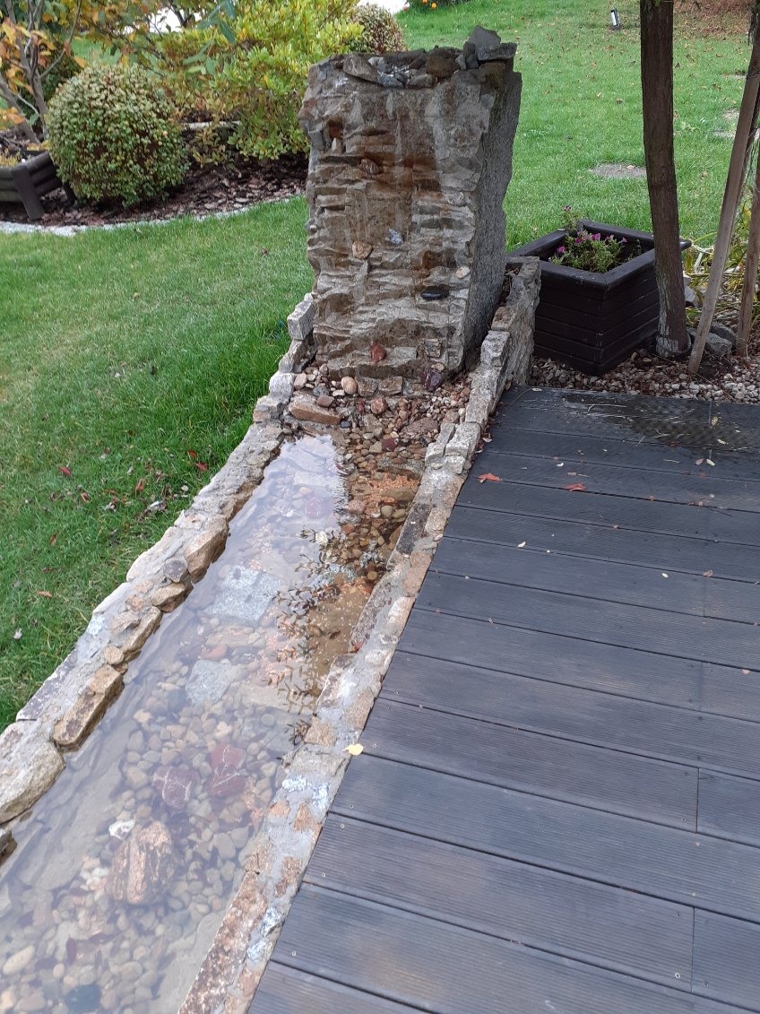 Oczko wodne z kamienia i drewniany taras we Wrocławiu. Kaskada z kamieni, woda spływa po kamieniach do koryta. Obok donica z kwiatami i trawnik.