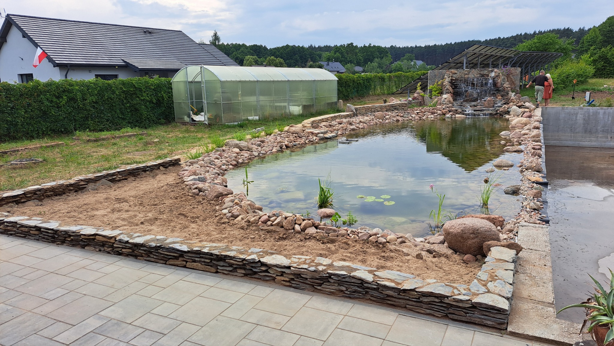 Oczko wodne w trakcie budowy z kamienną kaskadą i roślinami wodnymi, otoczone kamieniami i piaskiem. W tle szklarnia i dom z polską flagą.