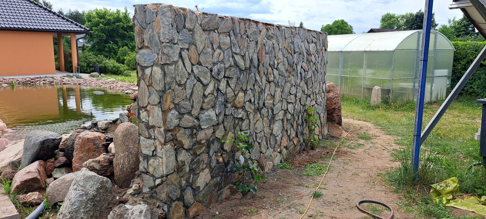 Kamienny mur oddzielający oczko wodne od ścieżki w ogrodzie, z widoczną szklarnią w tle. Wykonanie z nieregularnych kamieni w odcieniach szarości i brązu.