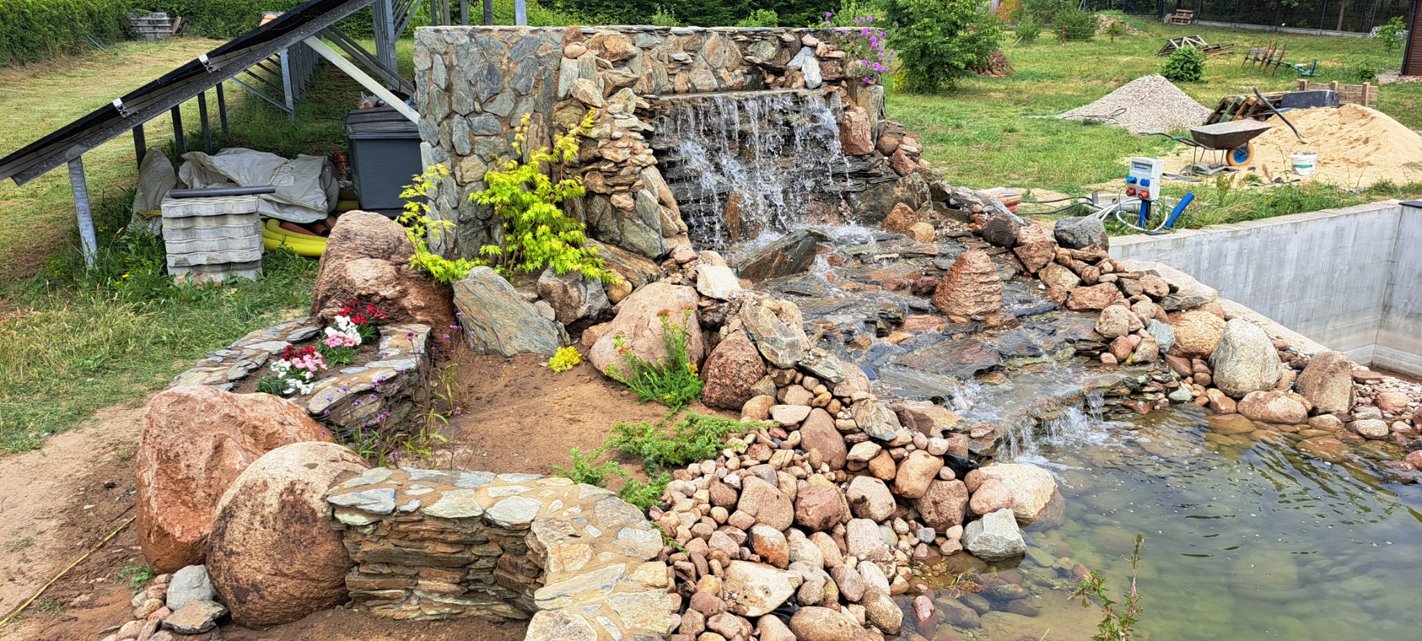 Oczko wodne z kaskadą, wykończone kamieniem naturalnym i zielenią. W tle widoczne panele słoneczne i materiały budowlane, sugerujące świeżo zakończone prace.