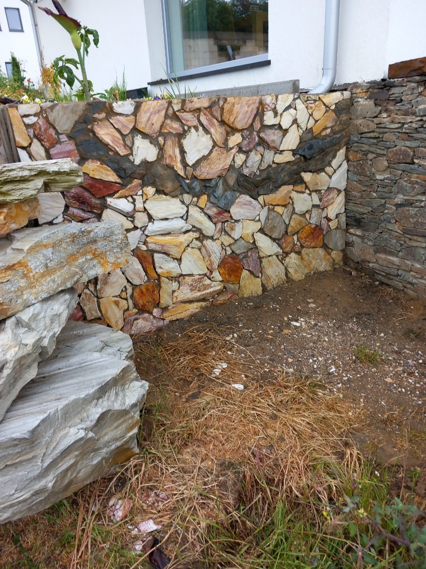Kamienny murek oporowy z nieregularnych, kolorowych płyt. Widoczne fragmenty ogrodu i elewacji budynku. Wykończenie z kamienia w odcieniach beżu, brązu i szarości.