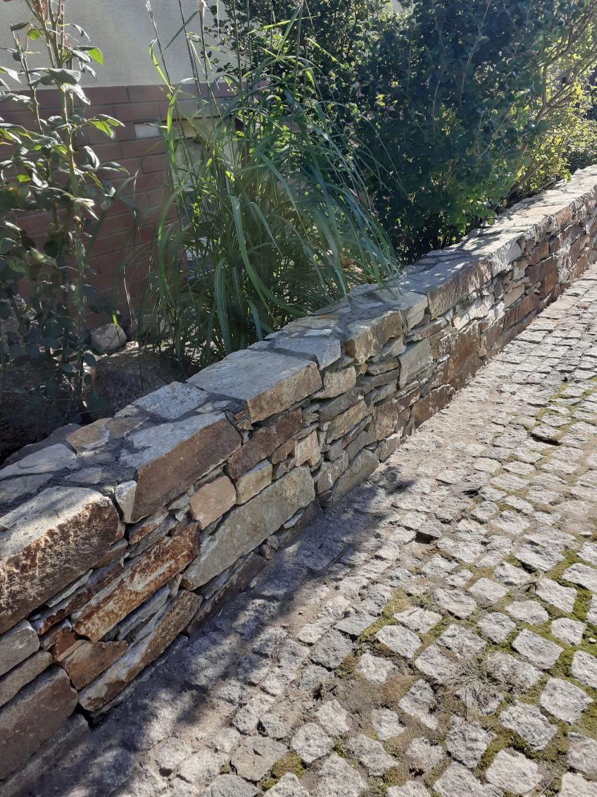 Kamienny murek oddzielający rabatę od ścieżki z kostki granitowej, z roślinnością w tle. Widoczne nieregularne ułożenie kamieni i cień na kostce.