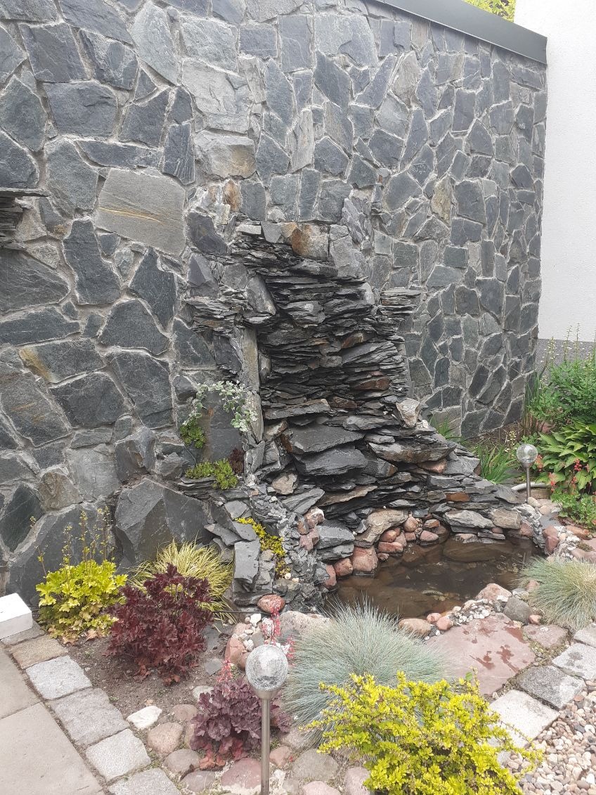 Oczko wodne z kaskadą z łupka na tle ściany z nieregularnego kamienia. Wokół rośliny ozdobne i ścieżka z kostki. Oświetlenie punktowe.