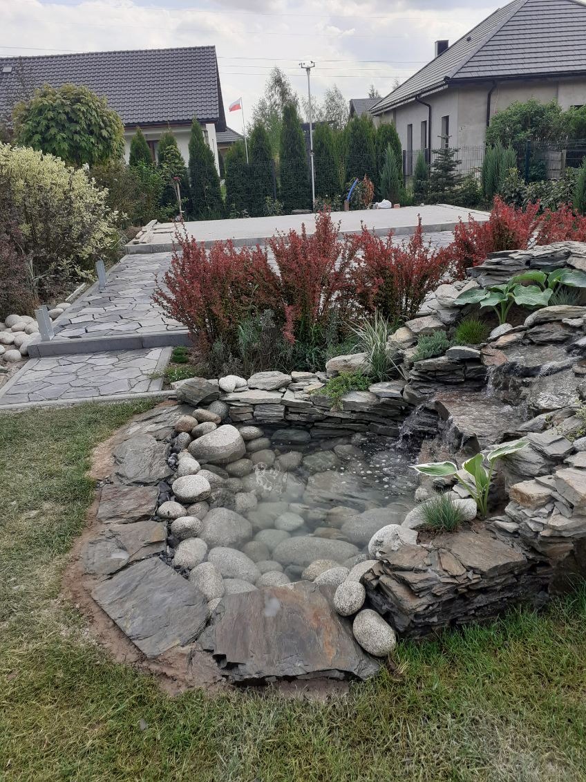 Oczko wodne z kaskadą wodną, obłożone kamieniami i roślinami. W tle widoczny kamienny chodnik i zadbany ogród z krzewami. Całość tworzy harmonijny krajobraz.