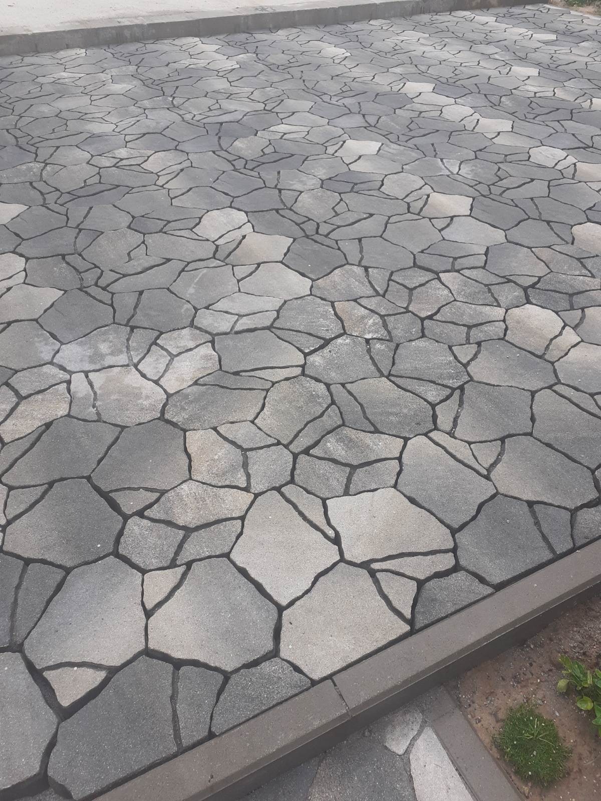 Ułożona nawierzchnia z nieregularnych, szarych płyt kamiennych, obramowana betonowym obrzeżem. Widok z góry, zieleń w rogu kadru. Nierównomierne odcienie szarości.