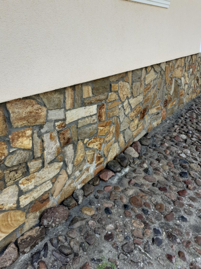 Kamienna elewacja domu z nieregularnych, brązowo-beżowych płytek. Podłoże wyłożone okrągłymi kamieniami. Detal wykończenia domu z bliska.