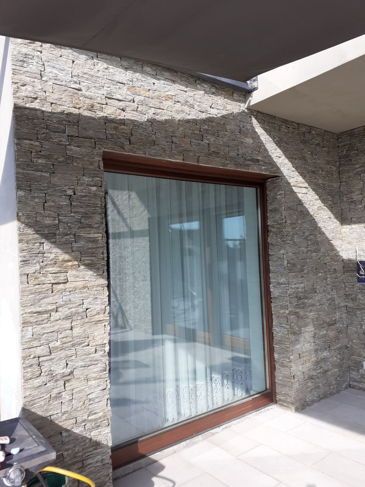Ściana zewnętrzna wyłożona dekoracyjnym kamieniem elewacyjnym w odcieniach szarości i beżu, z oknem w brązowej ramie. Widoczne fragmenty tarasu i zadaszenia.