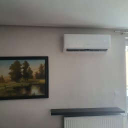 LUXIMAGO SPÓŁKA Z OGRANICZONĄ ODPOWIEDZIALNOŚCIĄ - Biała klimatyzacja ścienna z wyświetlaczem temperatury (24°C) w jasnym wnętrzu, obok obraz w czarnej ramie. Widoczny grzejnik i wyjście na balkon.