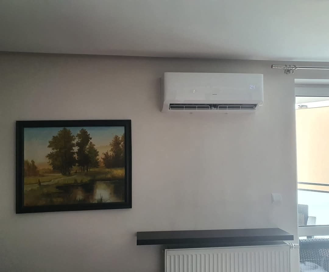 Biała klimatyzacja ścienna z wyświetlaczem temperatury (24°C) w jasnym wnętrzu, obok obraz w czarnej ramie. Widoczny grzejnik i wyjście na balkon.