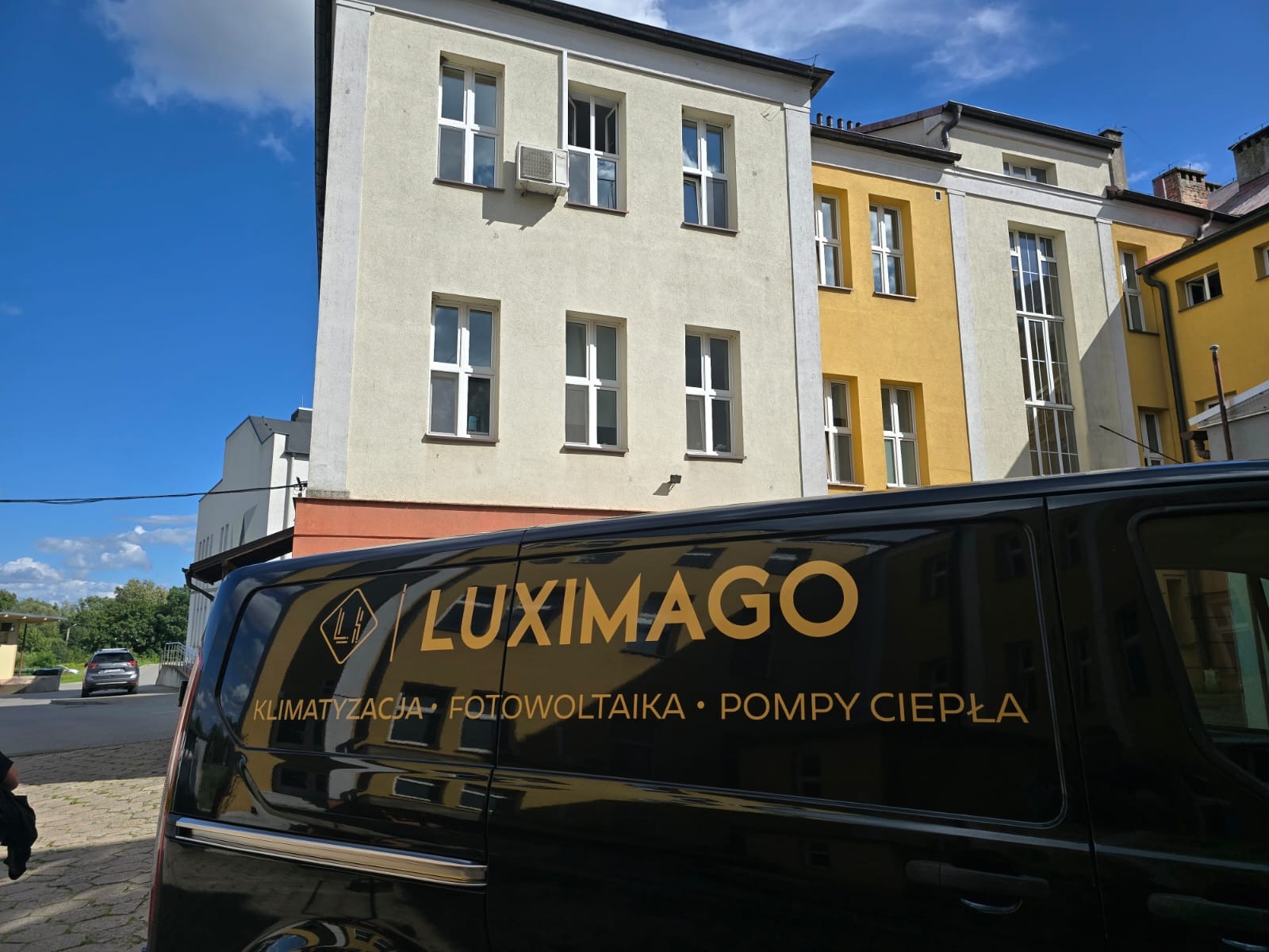 Czarny van firmy Luximago z logo i napisem: klimatyzacja, fotowoltaika, pompy ciepła na tle budynku z klimatyzatorem na ścianie. Błękitne niebo.