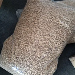 Dwa worki wypełnione pelletem drzewnym na szarej macie. Widoczne pojedyncze granulki i przezroczyste worki foliowe. Częściowo widoczny drewniany element.