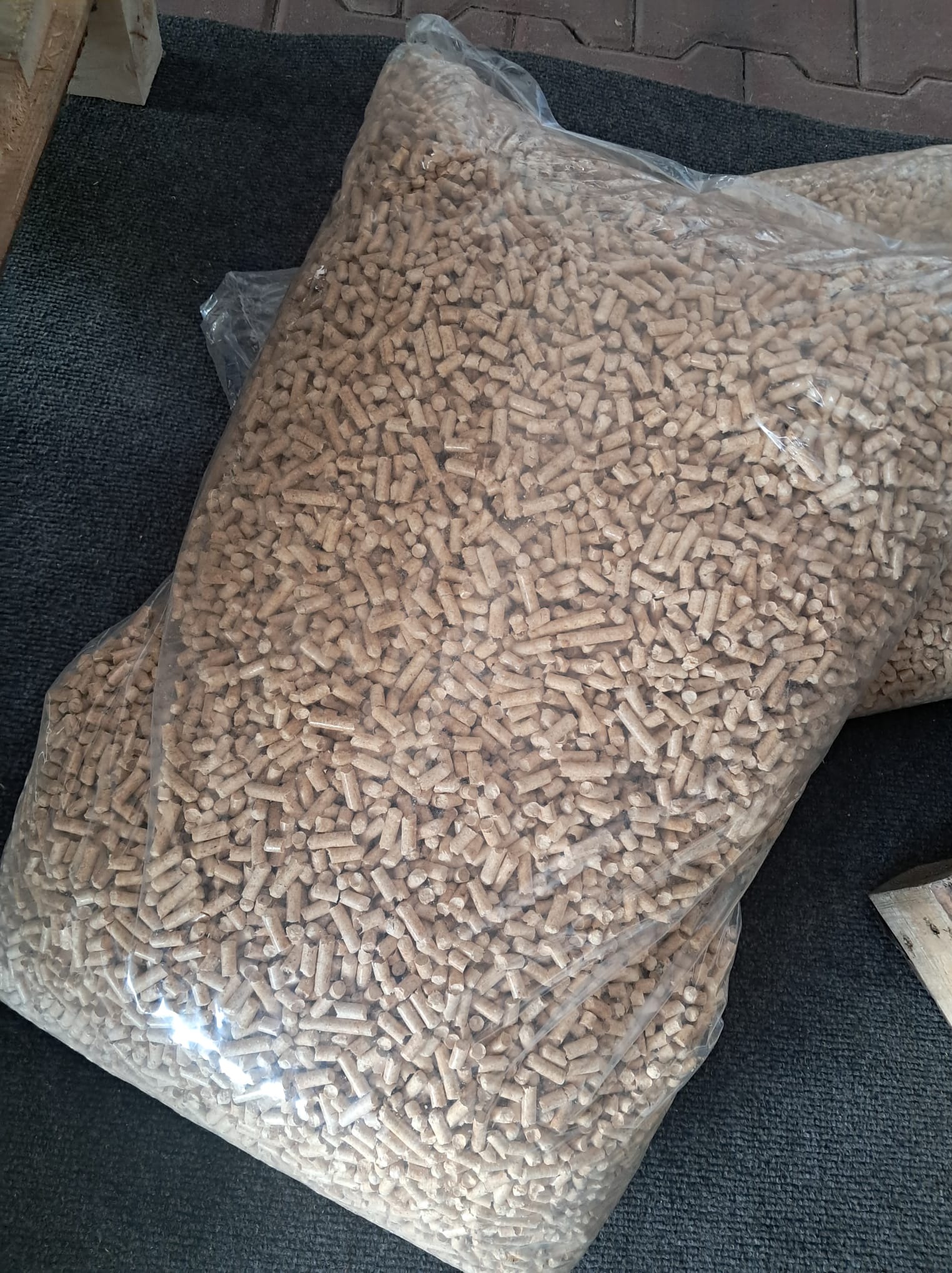 Dwa worki wypełnione pelletem drzewnym na szarej macie. Widoczne pojedyncze granulki i przezroczyste worki foliowe. Częściowo widoczny drewniany element.
