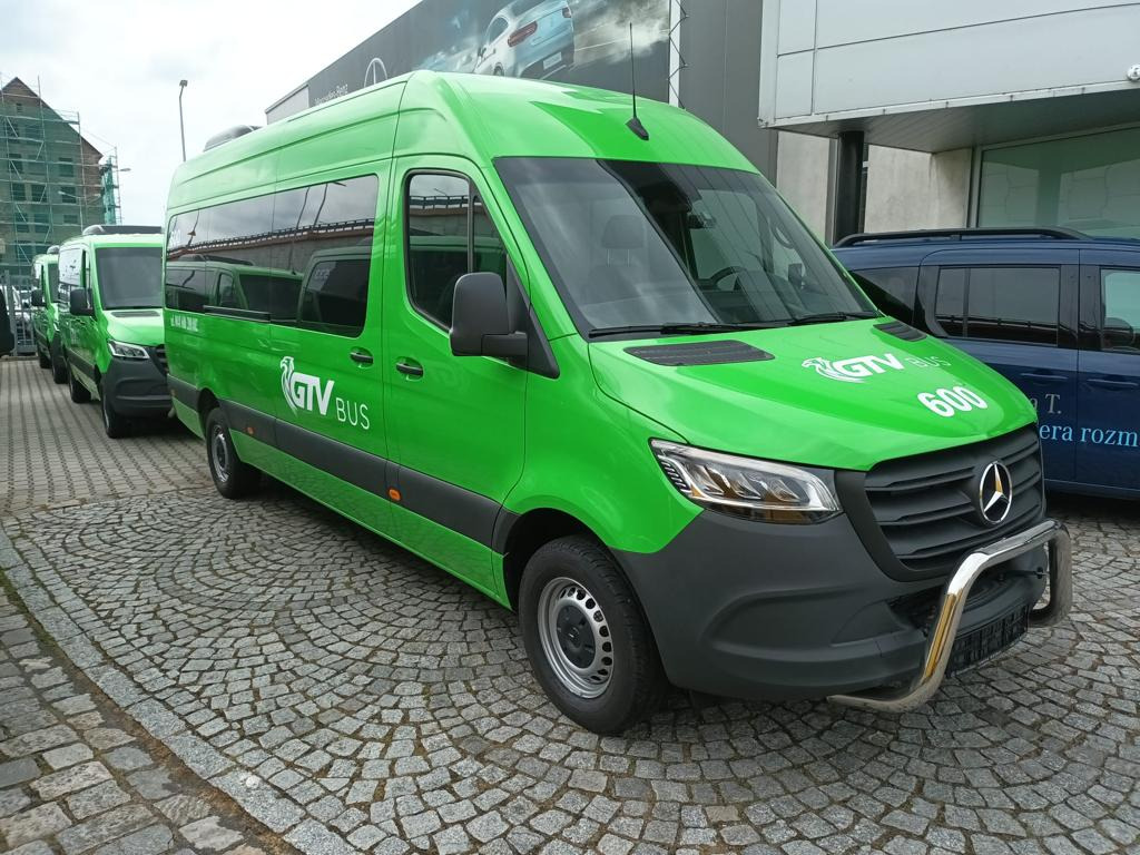 Szereg identycznych, zielonych busów GTV BUS na brukowanym placu, z metalowym orurowaniem z przodu. W tle budynek i reklama Mercedes-Benz.