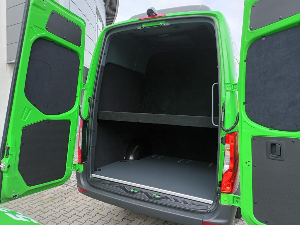 Wnętrze zielonego busa transportowego z otwartymi drzwiami, widoczna pusta przestrzeń ładunkowa z półką i zabezpieczeniami ścian.