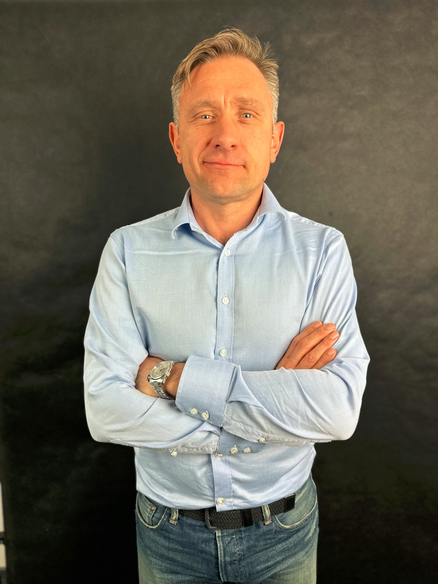 Portret mężczyzny w błękitnej koszuli z założonymi rękami na tle czerni, z widocznym zegarkiem na nadgarstku i delikatnym uśmiechem.