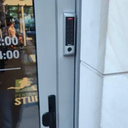 Szary panel z klawiaturą numeryczną, zamontowany na aluminiowej ramie drzwi wejściowych do studia treningowego, z widocznym logotypem ZKTeco i czarną klamką.