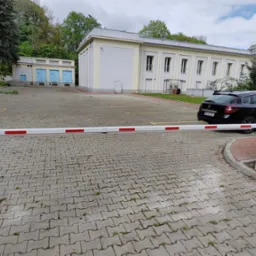 Biało-czerwona szlaban automatyczny blokuje wjazd na parking z czarnym autem i budynkami w tle. Widoczny panel sterowania szlabanem.