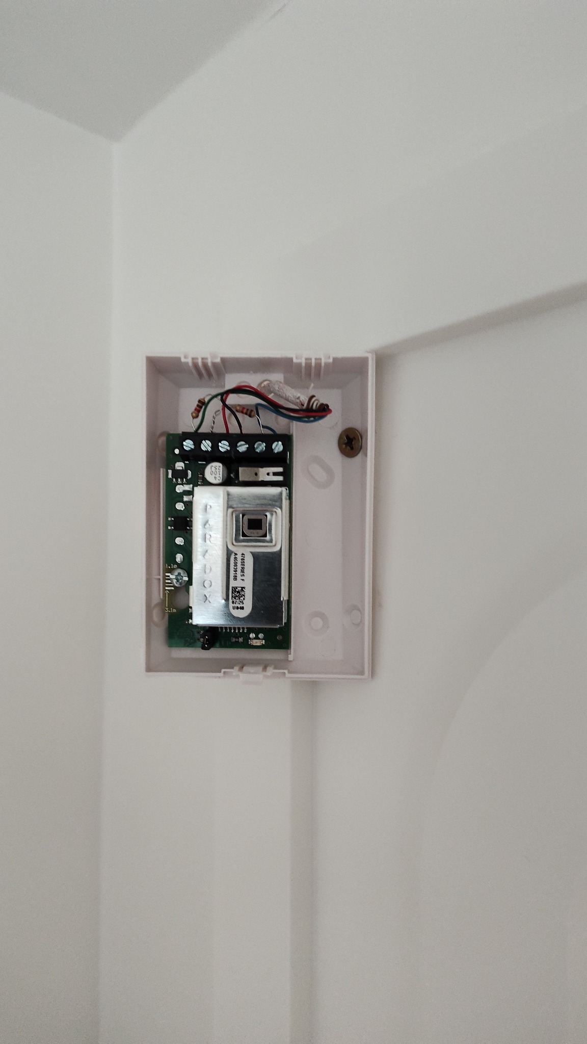 Otwarta obudowa czujnika alarmowego Paradox na białej ścianie. Widoczne przewody i elektronika. Śruba mocująca w prawym górnym rogu.
