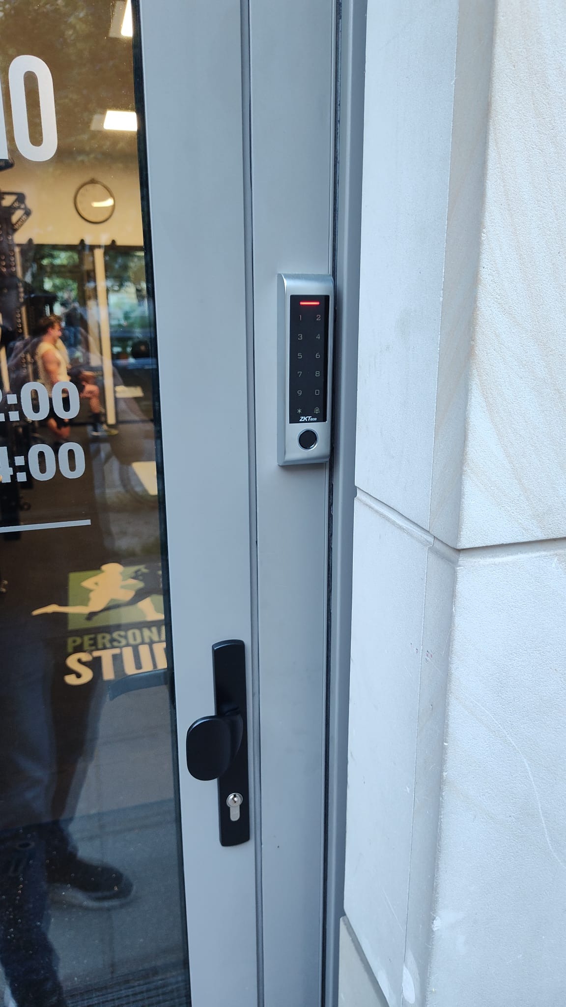 Szary panel z klawiaturą numeryczną, zamontowany na aluminiowej ramie drzwi wejściowych do studia treningowego, z widocznym logotypem ZKTeco i czarną klamką.
