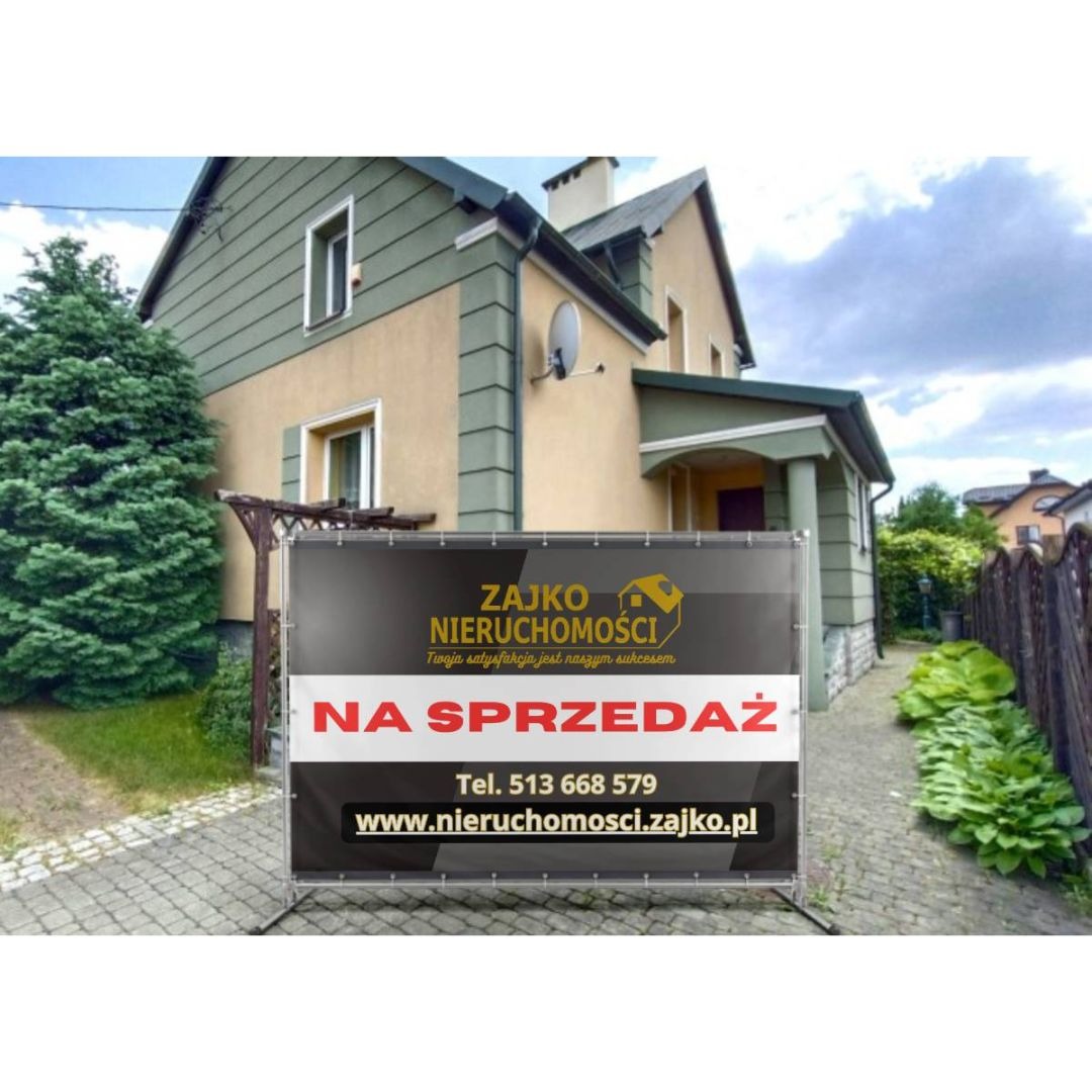 Baner 'Na Sprzedaż' firmy Zajko Nieruchomości na tle domu z ogrodem. Czarno-złoty baner z danymi kontaktowymi. Ujęcie z poziomu chodnika.
