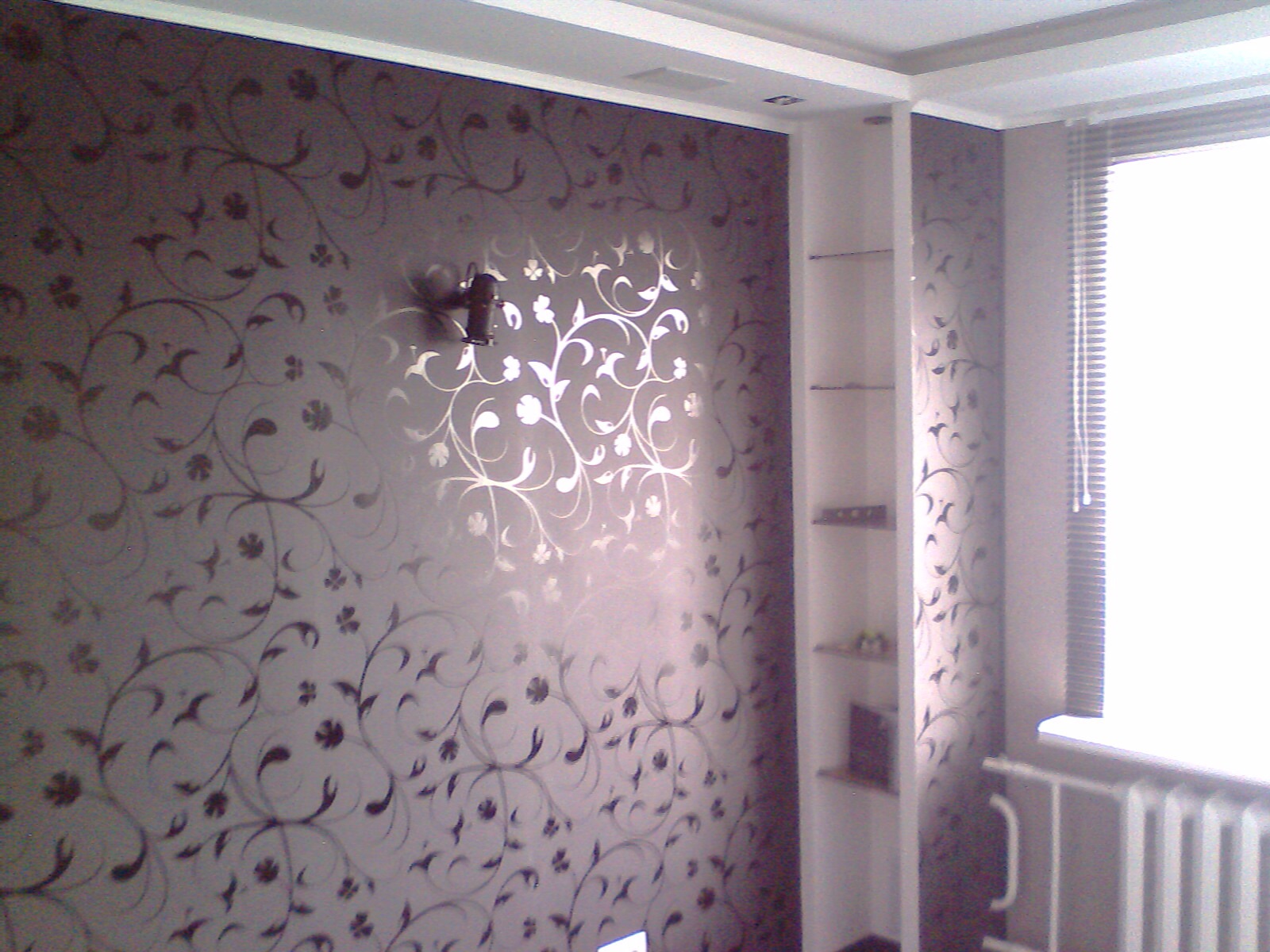 Ściana z wzorzystą tapetą w odcieniach brązu i srebra, z zamontowanym uchwytem na telewizor. Obok biała wnęka z półkami i okno z żaluzjami.