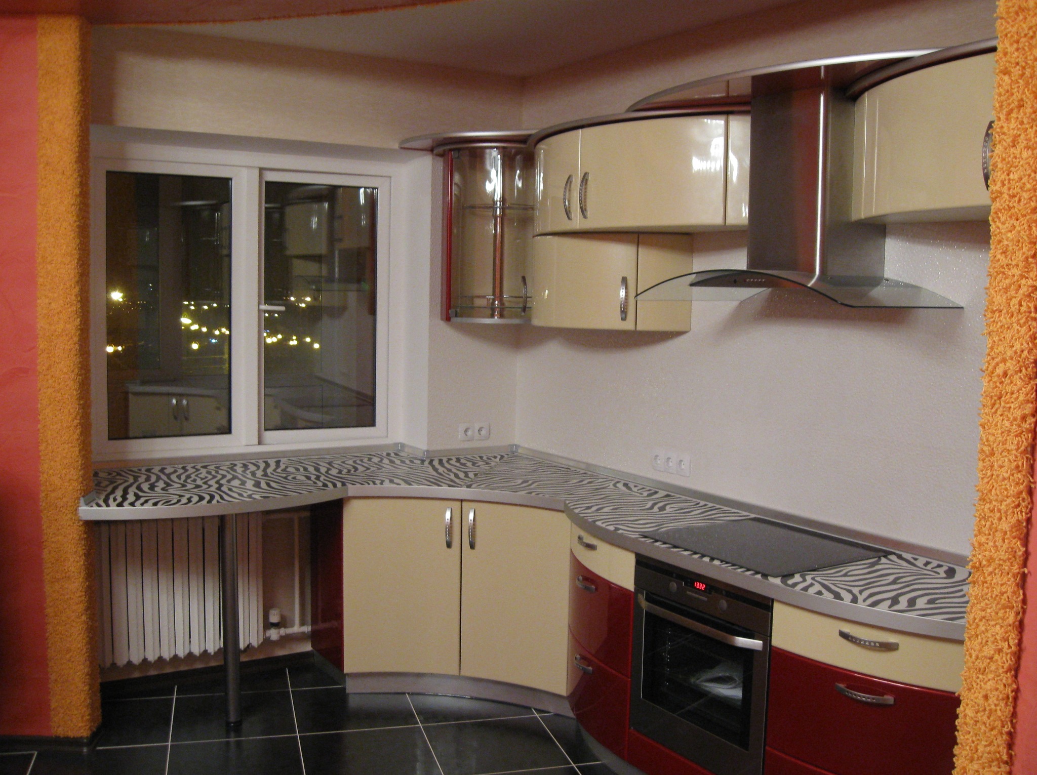 Ukończona kuchnia z blatem w zebrę, beżowymi szafkami i czerwonymi akcentami. Widok na okno z nocnym widokiem miasta. Nietypowe wykończenie.