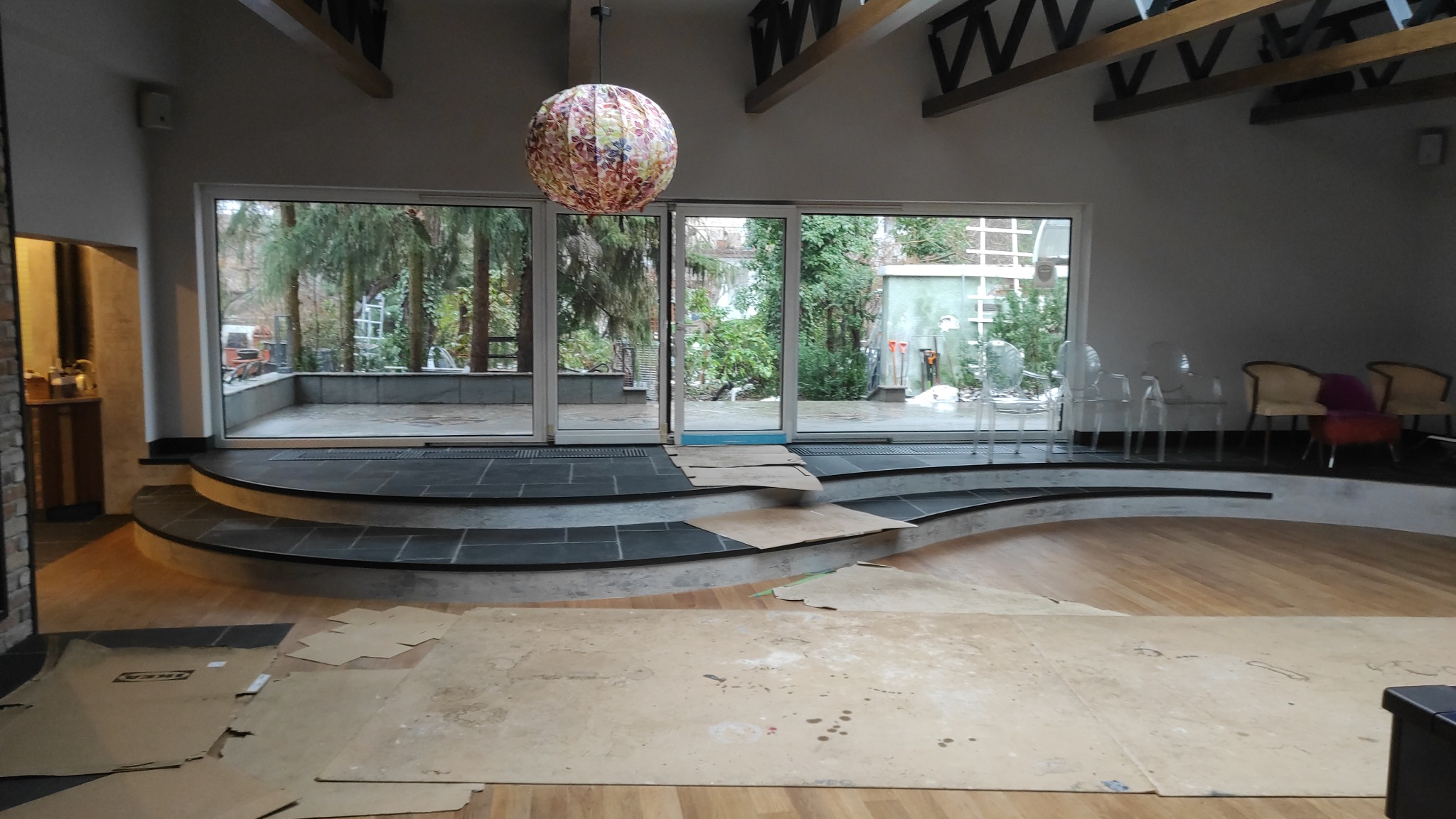 Wnętrze w trakcie prac wykończeniowych: zabezpieczona podłoga, schody z ciemnej terakoty, duże okna z widokiem na zieleń, dekoracyjna lampa wisząca.