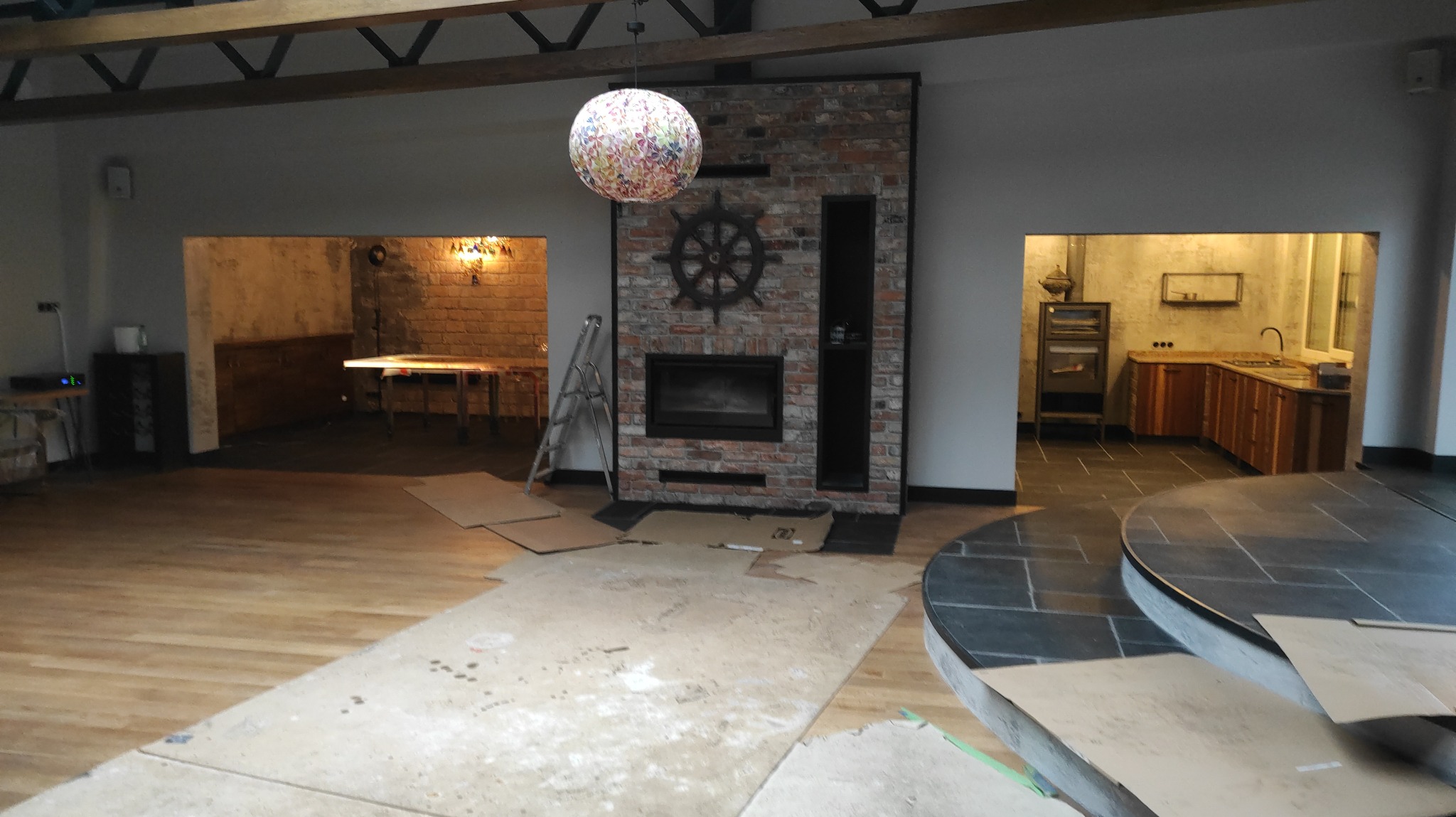 Wnętrze w trakcie prac: drewniana podłoga, ceglany kominek z kołem sterowym, kuchnia z drewnianymi szafkami w oddzielnym pomieszczeniu. Widoczny bałagan i zabezpieczenia.