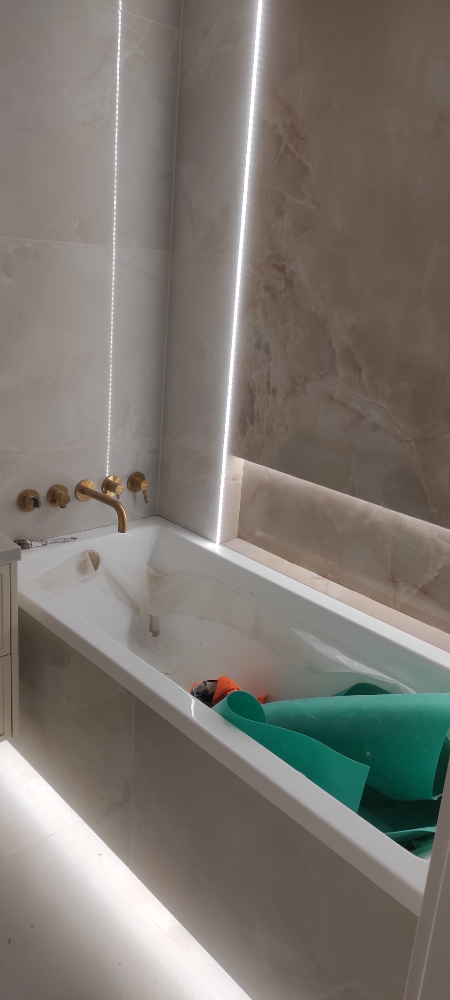 Biała wanna z złotą armaturą i podświetleniem LED na tle marmurowych płytek. W wannie zabezpieczenia malarskie. Detal wykończenia łazienki.