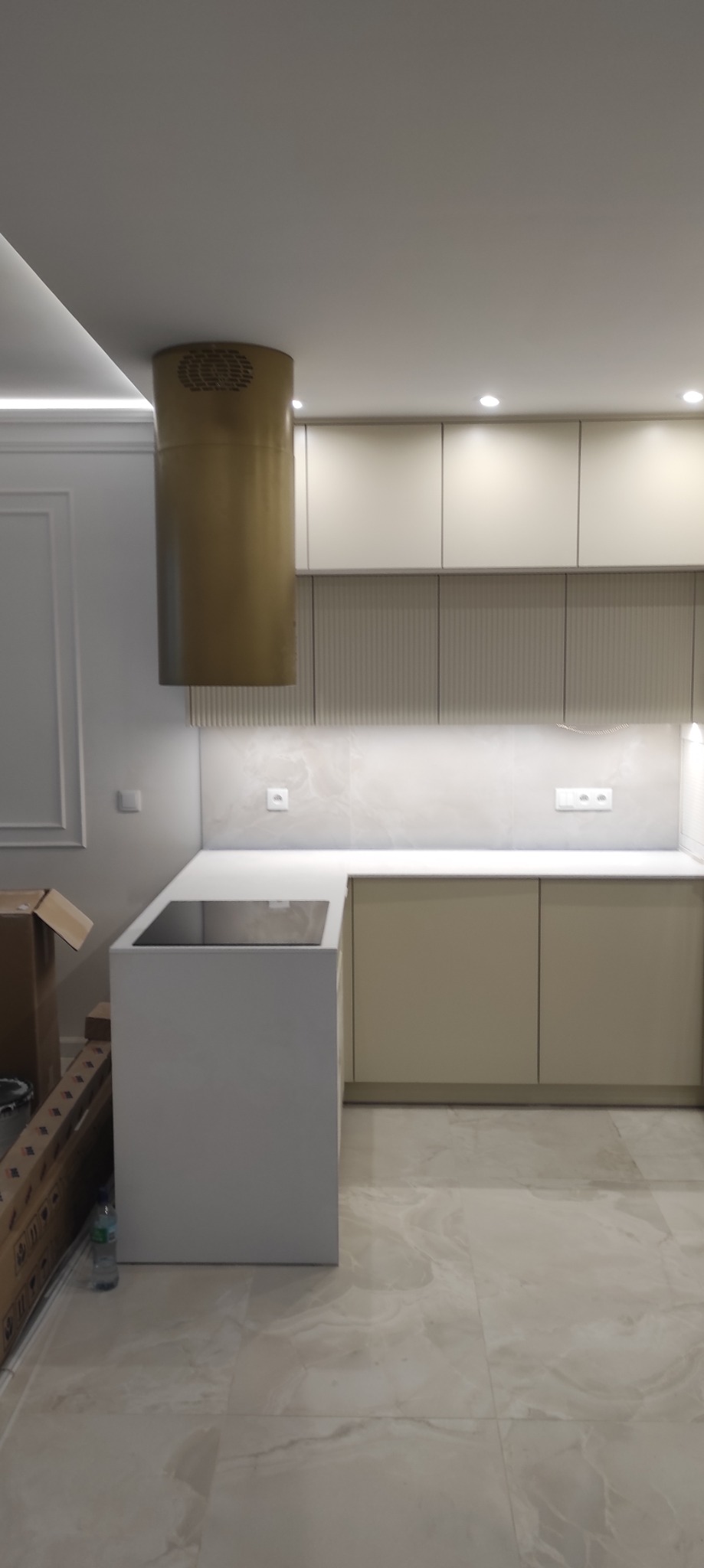 Nowoczesna kuchnia w odcieniach beżu z cylindrycznym okapem w kolorze złota, minimalistycznymi szafkami i marmurową podłogą. Częściowo widoczne kartony po lewej.