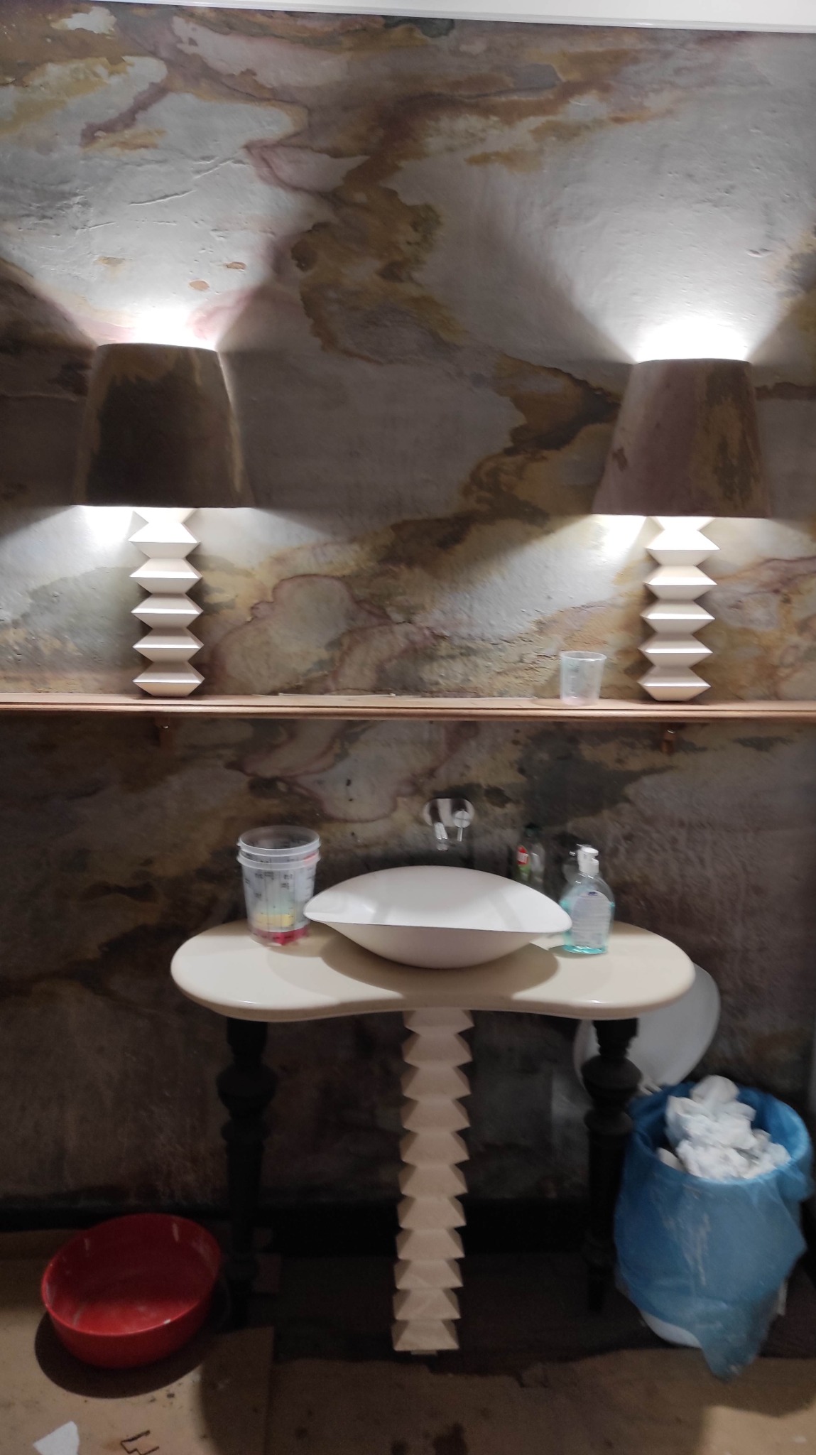 Wykończona łazienka z umywalką na dekoracyjnym stole, lampami ściennymi i artystycznym malowaniem sufitu. Widoczne akcesoria łazienkowe i wiadro z odpadami.