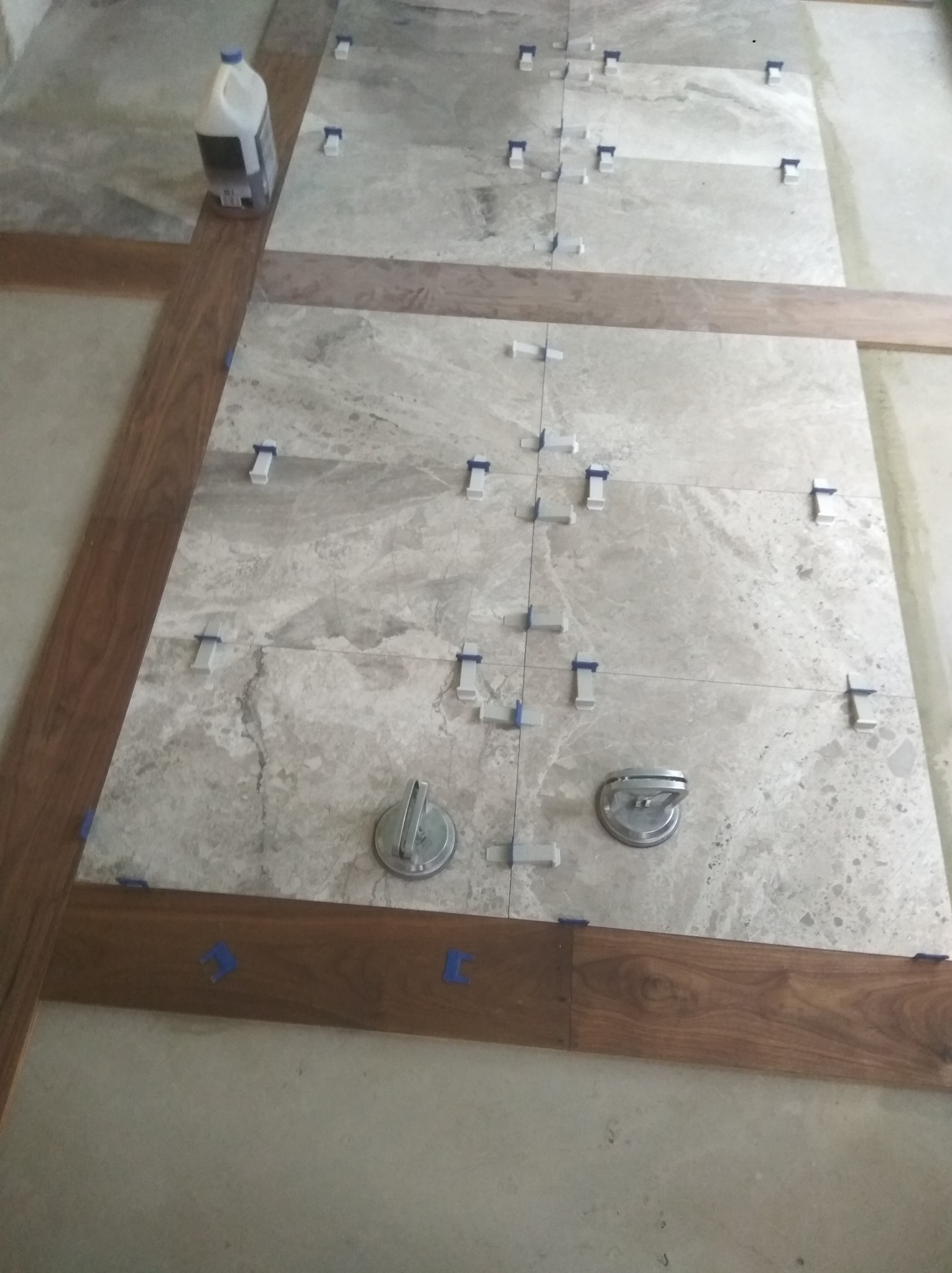 Układanie płytek podłogowych z użyciem systemów poziomowania i przyssawek. Widoczna butelka z płynem do czyszczenia. Prace wykończeniowe w trakcie realizacji.