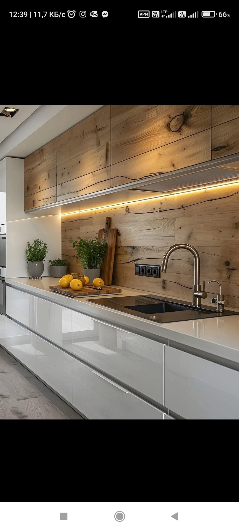 Nowoczesna kuchnia z białymi szafkami, drewnianą ścianą i oświetleniem LED. Na blacie cytryny, zioła w doniczkach i deska do krojenia. Minimalistyczny design.